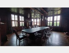 Holzvertäfelter Sitzungssaal mit einem langen ovalen Tisch und gepolsterten Stühlen. Große Fenster mit Sprossen und Glasornamenten lassen Tageslicht herein, und mehrere hängende Lampen beleuchten den Raum.