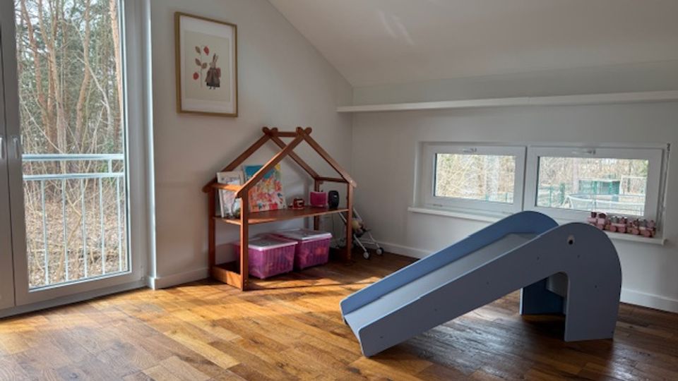 Bright attic playroom with a sloped ceiling, hardwood floor, and large windows and a glass door overlooking trees. The room includes a small indoor slide and a wooden house-shaped shelf with storage bins against a light-colored wall.