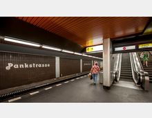 U-Bahn-Station „Pankstraße“ mit dunklen Fliesenwänden und weißer Beschriftung des Stationsnamens. Zu sehen sind ein Bahnsteig, Rolltreppen mit gelber Ausschilderung zum Ausgang sowie eine beleuchtete Decke mit orangen Paneelen.