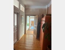 Ein Flur mit Holzboden und weißer Decke, auf der linken Seite eine Tür mit Glaseinsätzen und ein an der Wand hängendes Bild. Rechts steht ein roter Schrank und eine grüne Zimmerpflanze. Im Hintergrund öffnet sich eine geöffnete Holztür zu einem Raum mit weiteren Holzmöbeln und einer Badewanne.