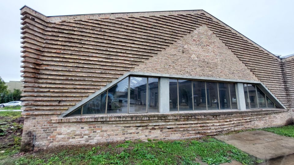 Ein modernes Backsteingebäude mit einer markanten, gestuften Fassadengestaltung aus vorspringenden Steinlagen. Ein großes, trapezförmiges Fenster mit Metallrahmen ermöglicht Einblick in den Innenraum; der umgebende Bereich ist mit Rasen und Gehwegplatten versehen.