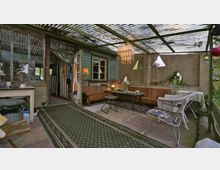 Überdachte Terrasse mit einem rustikalen Holzrahmen und Wellblechdach, ausgestattet mit einer Sitzecke aus lederbezogenen Sofas, einem kleinen Tisch und verschiedenen Stühlen. Die Wände sind teils aus verputztem Beton mit dreieckigen Fensteröffnungen, und der Boden ist mit Teppichen und Steinfliesen ausgelegt; im Hintergrund ein Bereich mit einem Küchentisch und einer offenen Tür.