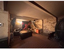 Industrieähnlicher Raum mit freiliegenden Ziegelwänden, Gewölbedecke und rotbraunem Fliesenboden. Im Vordergrund steht eine Sitzgruppe mit dunklen Ledersofas um einen Holztisch, im Hintergrund befinden sich ein großes rotes "X", eine silberne Rutsche sowie verschiedene Möbel und Lampen.