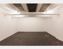 Leerer Innenraum mit weißen Wänden, dunklem Fliesenboden und einer sichtbaren Betondecke mit quer verlaufenden weißen Balken und einfachen Deckenleuchten.