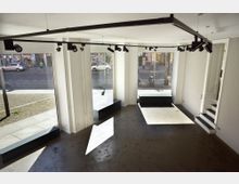 Innenraum eines leeren, modernen Ladenlokals mit großen Schaufenstern, durch die eine Straße mit parkenden Autos und Fahrrädern sichtbar ist. Der Raum hat weiße Wände, dunklen Boden und eine schwarze Deckenkonstruktion mit Strahlern, sowie Stufen im hinteren Bereich.