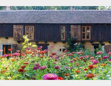 Ein ländliches Gebäude mit dunkler Holzfassade und geöffneten Fensterläden auf einem gelben Putzsockel, umgeben von einem Garten mit bunten Blumen im Vordergrund. Im Hintergrund sind Bäume zu sehen.