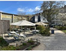 Ein kleiner Außenbereich eines Cafés mit gepflastertem Boden, weißen Tischen, grünen Klappstühlen und großen Sonnenschirmen. Im Hintergrund ist ein Gebäude mit schiefergedecktem Dach sowie blühende Bäume in einer parkähnlichen Umgebung zu sehen.