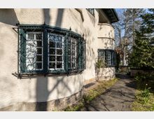 Teilansicht eines historischen Wohnhauses mit cremefarbener Putzfassade und grünen, verzierten Gitterfenstern. Im Vordergrund verläuft ein gepflasterter Weg, umgeben von Gartenbewuchs und Bäumen.