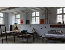 Ein heller Werkraum mit hohen Fenstern und mehreren Holztischen, die mit Werkzeugen, Modellen und Materialien belegt sind. An den Wänden befinden sich künstlerische Plakate, geometrische Rahmen und Regale mit weiteren Utensilien.