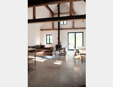 Offener Wohnraum mit hohen Decken und sichtbaren Holzbalken, ausgestattet mit einem Sofa, einem Kaminofen und einem langen Esstisch. Große Fenster und Türen sorgen für viel Tageslicht, der Boden besteht aus poliertem Beton.