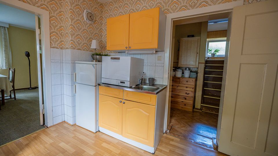Kleine, ältere Küche mit gelben Hängeschränken, weißen Fliesen an der Wand, Kühlschrank, Spüle und einer kompakten Spülmaschine auf der Arbeitsfläche. Gemusterte Tapete im Retro-Stil, Laminatboden und offene Türdurchgänge zu angrenzenden Räumen.