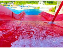 Rote Wasserrutsche in einem Freibad, aus der Wasser sprudelt, mit Blick auf ein großes Außenbecken, beige Pflasterflächen und umliegende grüne Vegetation.