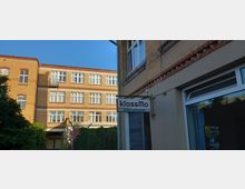 Backsteinfassaden eines historischen Gebäudekomplexes mit großen weißen Sprossenfenstern. Im Vordergrund ein Schild mit der Aufschrift „klassMo Kulturwerkstatt“ vor einem Eingangsbereich, umgeben von Bäumen und Efeu.