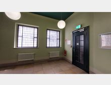 Ein Flur mit grün gestrichenen Wänden, zwei großen Fenstern mit Gittermustern und einem schwarzen Türrahmen mit Glaseinsätzen. An der Wand befinden sich Heizkörper, ein Feuerlöscher, ein beleuchtetes Notausgangsschild und ein Flucht- und Rettungsplan.