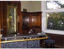 Kleine Bar mit dunkelbrauner Holzvertäfelung, einer schwarzen Granittheke und einem Kühlschrank mit beleuchteten Getränken. Rechts ein großes Fenster mit Blick auf Bäume und ein Heizkörper unter der Fensterbank.