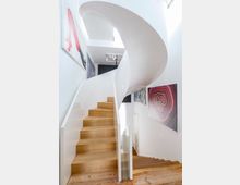 Moderne, helle Treppe mit einer skulpturalen, weißen Wendung und Holzstufen, umgeben von weiß gestrichenen Wänden mit abstrakten Kunstwerken. Oberhalb der Treppe ist ein Deckenausschnitt mit zusätzlichem Licht und einer kleinen Deckenleuchte zu sehen.