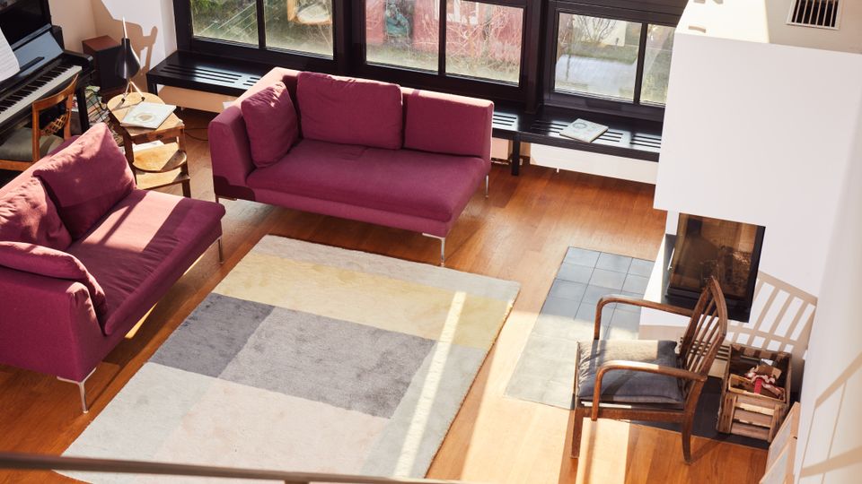 A modern living room with large floor-to-ceiling grid-patterned windows, allowing natural light to fill the space. The room features wooden flooring, a pair of magenta sofas, a patterned rug, and a contemporary fireplace near a wooden chair, viewed from the top of a staircase with metal railings.