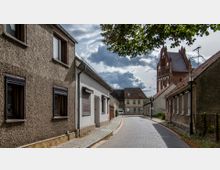 Schmale gepflasterte Straße in einer ruhigen Wohngegend mit einfachen Häuserfassaden auf beiden Seiten. Im Hintergrund ragt eine Backsteinkirche mit markanten Ziergiebeln empor, während einige Wolken den Himmel bedecken.