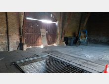 Innenansicht eines alten, verlassenen Gebäudes mit freiliegenden Ziegelwänden. Zu sehen sind ein großes hölzernes Tor, ein Loch im Boden mit einem Metallgitter, Mülltonnen und eine ältere, blauüberdachte Rikscha.