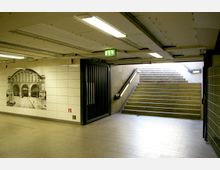 Unterirdischer Übergang mit Fliesenwänden, auf denen ein historisches Bild eines Bahnhofs abgebildet ist. Rechts führen eine beleuchtete Treppe und ein Geländer nach oben ins Freie, während die Decke mit einfachen Lampen beleuchtet ist.
