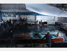 Innenraum eines Filmstudios mit einem großen Wasserbecken, in dem mehrere Personen an einem Filmset arbeiten. Im Vordergrund liegt ein elegantes Holzmotorboot am Beckenrand, während im Hintergrund technische Ausrüstung und Crewmitglieder sichtbar sind.