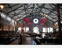 Großzügiger Innenraum eines Restaurants in einer ehemaligen Industriehalle mit Stahlträgerdecke, gemauerten Wänden und hohen Bogenfenstern. Die Rückwand ist mit einem bunten Wandgemälde verziert, das einen Kraken und mehrere Schiffe zeigt, und der Raum ist mit Holztischen und modernen Stühlen ausgestattet.