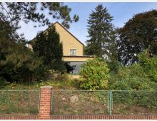 Zweistöckiges Wohnhaus mit gelber Fassade, umgeben von dichter Vegetation und Bäumen. Im Vordergrund eine Metallzaun-Konstruktion auf einem roten Backsteinsockel.