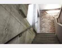 Betontreppe in einem industriellen Innenraum, mit roher Ziegelwand und sichtbaren Betonwänden. Eine Metallhandlauf verläuft entlang der linken Wand, und eine Deckenleuchte beleuchtet den Bereich.
