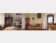 Ein historischer Raum mit Holzboden, antiken Möbeln und gemusterten Tapeten. Links steht ein gläserner Vitrinenschrank, in der Mitte ein runder Esstisch mit Stühlen, und rechts führen geöffnete Türen in angrenzende Räume mit hohen Fenstern und weiteren klassischen Elementen.