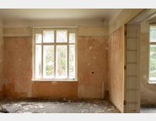 Innenraum eines leerstehenden, renovierungsbedürftigen Zimmers mit großem, weiß gerahmtem Fenster und abgenutzten, teils beschädigten Putzwänden. Rechts führt ein breiter Durchgang in einen weiteren Raum mit zusätzlichem Fenster; auf dem Boden liegen Schutt und abblätternde Materialien.