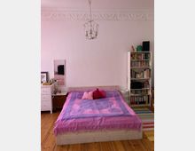 Ein Schlafzimmer mit Holzdielenboden, einem weißen Bett, das mit einer lila-rosa Tagesdecke und farbigen Kissen dekoriert ist. Im Hintergrund befinden sich ein Bücherregal, eine Kommode und eine Wand mit Stuckverzierungen sowie ein Kronleuchter an der Decke.