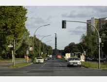 Straßenansicht in Berlin mit der Straße des 17. Juni, von der Bismarckstraße aus gesehen, die in Richtung der Siegessäule führt. Die Straße ist von Bäumen flankiert, gesäumt von Autos und Leuchtreklamen, während Straßenschilder und Ampeln am Rand sichtbar sind.