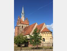 Backsteinkirche mit roten Dachziegeln und einem hohen Kirchturm mit Spitze und Kreuz, umgeben von hellem Himmel. Die Fassade besteht aus Feld- und Ziegelsteinen, und es gibt einen kleinen Baum sowie einen Metallzaun im Vordergrund.