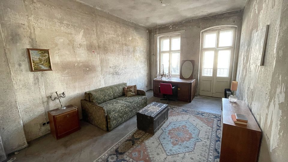 Großes, spärlich möbliertes Zimmer mit abgenutzten, unverputzten Wänden, einem gemusterten Sofa, Teppich und Holzmöbeln. Am Ende des Raums stehen zwei hohe Fenster mit einer verglasten Tür, die Tageslicht hereinlassen.