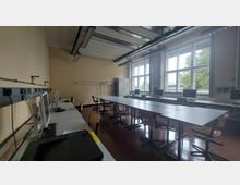 Ein Klassenraum mit mehreren Tischen und Stühlen, ausgestattet mit Computern entlang der Wand. Große Fenster lassen Tageslicht einfallen, und an der Decke sind technische Beleuchtungselemente montiert.