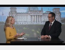 Eine Frau und ein Mann stehen an einem Stehtisch und sprechen miteinander, im Hintergrund ist eine großflächige Projektion des Reichstagsgebäudes mit der Glaskuppel und dem Fernsehturm von Berlin sichtbar. Auf dem Tisch stehen zwei Gläser mit Wasser.