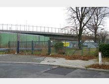 Ein eingezäuntes Areal mit Bäumen und Laub am Boden, im Hintergrund eine grüne Schallschutzwand und eine erhöhte Brücke mit Geländer. Im Vordergrund sind ein Gehweg, eine Straßenkante und ein weißer Pfosten erkennbar.