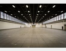 Weitläufige, leere Messehalle mit hohen weißen Wänden, einer dunklen Decke mit zahlreichen rechteckigen Deckenleuchten und großen Fenstern entlang der oberen Wandbereiche. Im Hintergrund befindet sich ein großer Eingang mit der Beschriftung "17".