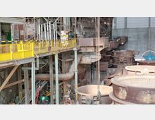 Industrieanlage im Innenbereich mit rostigen Metallstrukturen, gelb gestrichenen Sicherheitsgeländern und großen metallenen Behältern. Im Hintergrund sind Rohrleitungen, Maschinenteile und eine Betonwand zu sehen.