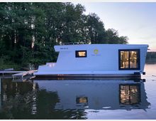 Ein modernes Hausboot mit weißer Außenwand und großen Fenstern schwimmt auf einem ruhigen See, umgeben von dichter Waldlandschaft. Im Inneren des Boots sind durch die beleuchteten Fenster Möbel und Dekorationen sichtbar.