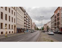 Straßenszene in einer Stadt mit mehrstöckigen Wohnhäusern auf beiden Seiten, Balkonen und Geschäften im Erdgeschoss; eine zweispurige Fahrbahn mit parkenden Autos, Gehwegen und einer Bushaltestelle auf der linken Seite unter bedecktem Himmel.