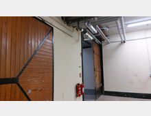 Ein schlichter Innenraum mit Holz-Schiebetüren an der Wand, sichtbaren Rohren an der Decke und zwei Feuerlöschern, die an einer hellen Wand angebracht sind. Der Raum wirkt wie ein Durchgangsbereich oder technischer Lagerraum.