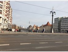 Straßenszene in Berlin mit Blick auf die Spree, die historischen Gebäude der Museumsinsel, den Berliner Fernsehturm im Hintergrund und eine verzierte Straßenlaterne im Vordergrund. Links befindet sich ein mehrstöckiges Wohngebäude, rechts moderne Bürogebäude und Gehwege mit Passanten und Fahrrädern.