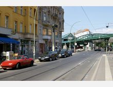 Straßenszene in einer Stadt mit Wohn- und Geschäftsgebäuden, darunter ein gelbes Gebäude mit Ladengeschäften im Erdgeschoss. Im Hintergrund ist eine grüne Hochbahnbrücke zu sehen, die über die Straße führt, mit Autos, geparkten Fahrzeugen und Straßenbahnschienen im Vordergrund.