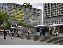 Ein städtischer Platz mit Marktständen auf einer breiten Treppe, umgeben von modernen Gebäuden. Links steht ein Bürogebäude mit großen Fenstern und bunten Firmen-Schildern, rechts eine graue Fassade mit geometrischen Mustern.