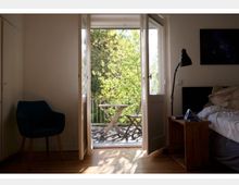 Ein Schlafzimmer mit Holzboden, einem blauen Sessel links und einem Bett rechts. Im Zentrum führen geöffnete Glastüren zu einem Balkon mit einem kleinen Tisch und zwei Stühlen, umgeben von Bäumen im Sonnenlicht.