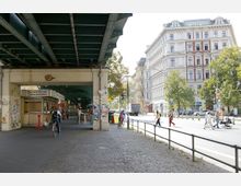 Straßenszene in einer Stadt mit erhöhten Eisenbahnschienen, die auf metallenen Trägern verlaufen. Unter den Schienen befinden sich ein kleiner Imbissstand und Fußgänger, während rechts der Straße historische Wohngebäude mit hellen Fassaden und Bäumen im Vordergrund zu sehen sind.