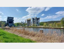 Industriearchitektur an einem Kanal mit grünem Ufer: Links befindet sich eine große, stählerne Hebekonstruktion, während rechts ein modernes Gebäude mit verglasten Fassaden und gelben Akzenten zu sehen ist. Im Hintergrund sind ein Hügel und ein bewaldetes Gebiet unter einem blau bewölkten Himmel zu erkennen.