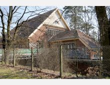 Backsteingebäude mit Satteldach in einem bewaldeten Umfeld, umgeben von einem Maschendrahtzaun. Das Gebäude zeigt Spuren von Graffiti, und im Vordergrund sind kahle Bäume sowie eine Rasenfläche sichtbar.