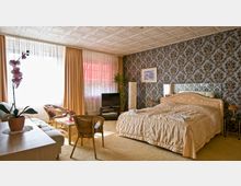 Ein gemütlich eingerichtetes Schlafzimmer mit einem großen Bett, einer floralen Tapete, einem Fernseher, einem Spiegel an der Wand und einem kleinen Sitzbereich mit Korbstühlen. Vorhänge bedecken teilweise die Fenster, durch die Tageslicht einfällt; eine Orchidee und weitere Pflanzen ergänzen die Dekoration.